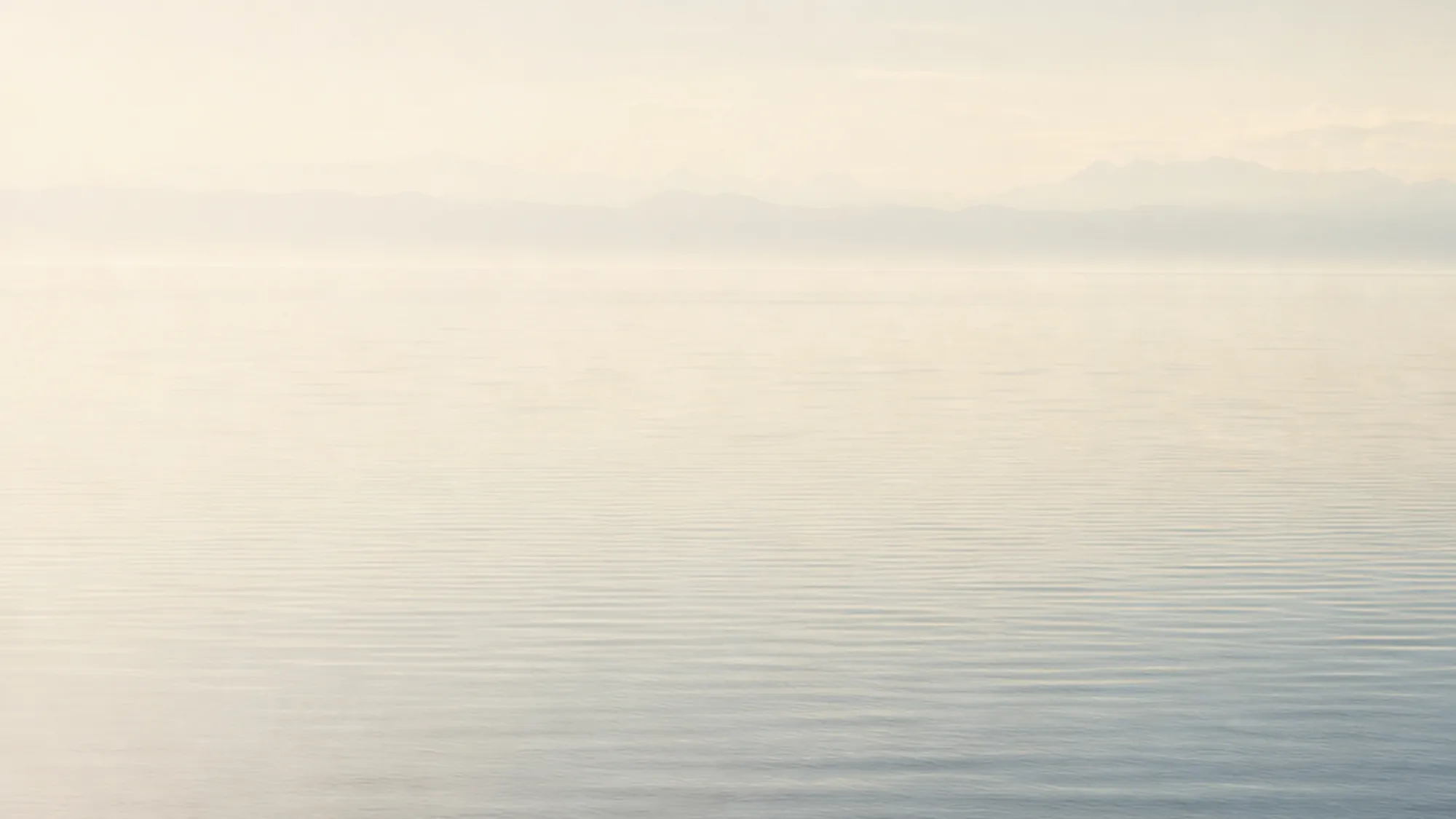 A calm water surface at early morning, gentle ripples catching pale daylight.